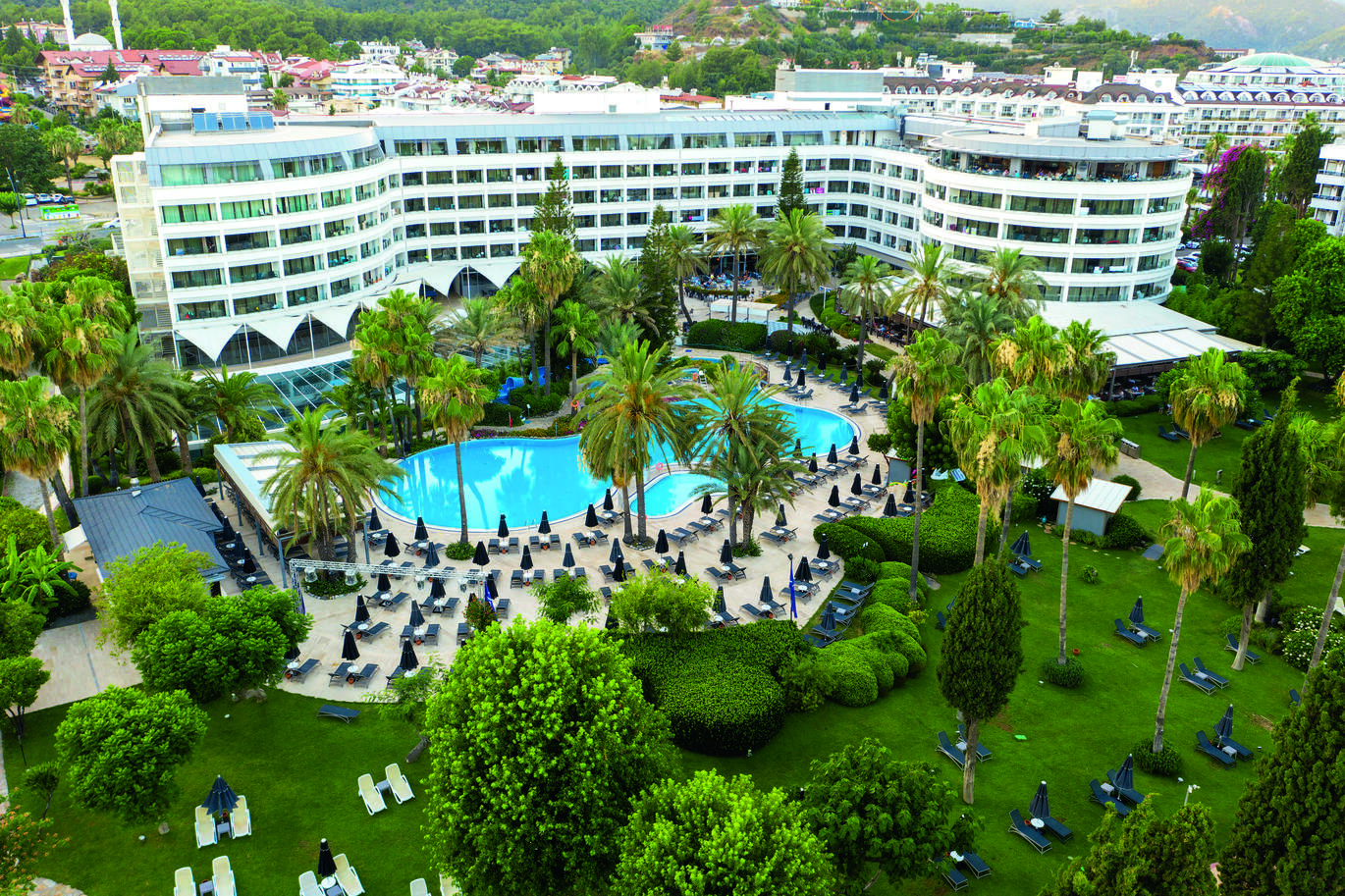 Large white hotel building with a cluster of rooms. Lots of green palm trees in front of building with a blue pool in front and lots of sun beds surrounding the pool. Sun beds also spill out onto the green lawn with a lot of trees and hedges around.
