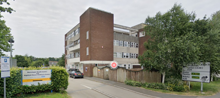 The hospital is shown along with signage to the left and right of the frame, and trees in the foregound and to the right of the hospital