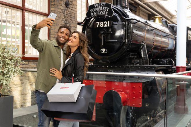 "Two people taking a selfie in front of a historic black steam locomotive labeled “West Somerset Railway 7821,” inside a building with brick walls and glass roof. They are holding multiple shopping bags"