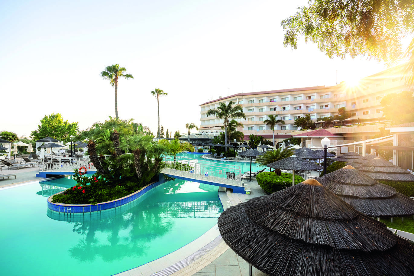View of the pool area with a small island in the middle and a bridge over leading to either side. With trees dotted around and the hotel building behind with the sun setting behind the building.