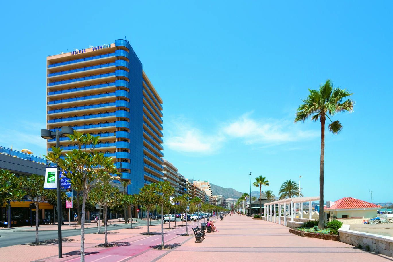 View of the tall hotel building from the street opposite lined with palm trees and mountains just visible in the background. 