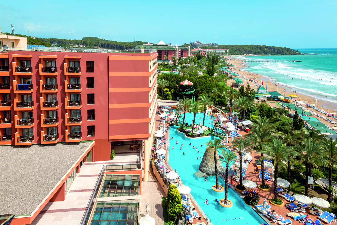A large dark red hotel building with a cluster of balconies on the side with a large pool below surrounded by many trees, blue sun loungers and white umbrellas with people in the pool and around it. There is a small bridge at the top end of the pool from one side to the other. To the right of the image is the sea and beach also with a number of sun loungers and many people on the beach and in the sea. There are other hotel buildings in the background and a forest of trees beyond with the clear blue sky above.