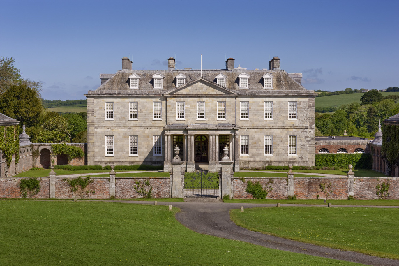 The south front of the house at Antony, Cornwall. The house was built between 1718 and 1724, and is faced with silver-grey Pentewan stone, with pedimented porte-cochere was added in the mid-nineteenth century. The building has a large pillared entrance with steps leading up to it and many many windows and chimneys. There is a brick wall surrounding the front garden of the building with some black metal gates in the centre. There is a driveway leading up to the gates through some gardens. There are many trees and some fields in the background with the blue sky above.