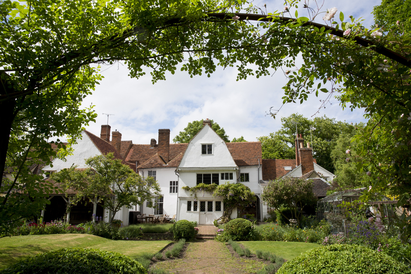 A view of the large white house through a vine archway with the lush green lawn filled with plants, flowers and greenery with a pathway through the middle.