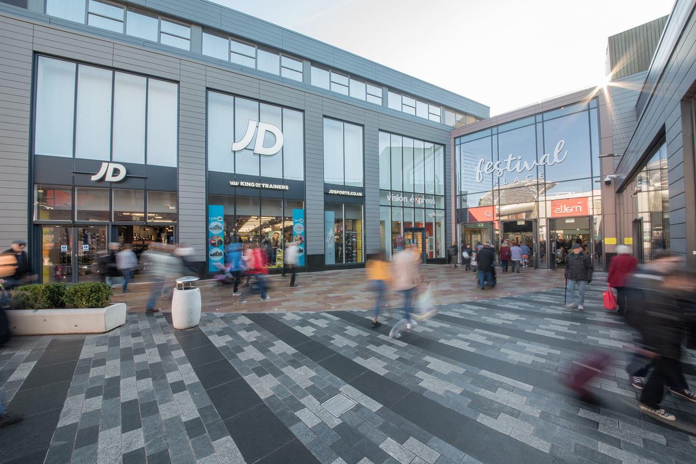 "Festival Place Entrance located to the right. Grey and white paving on route to the entrance. JD Sports located straight ahead. People are blurred in the image."