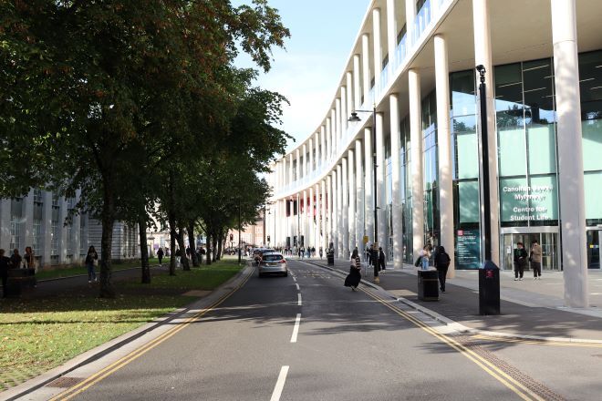 "Centre of Student Life to the right, with a road in front. Trees follow the road on the left, with students walking down each side"