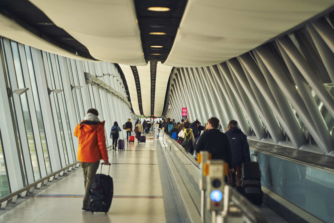 "People walking along a vast corridor pulling their luggage, with windows on either side, at London Gatwick North Terminal Arrivals"