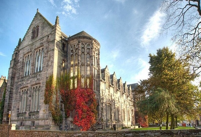 "External image of an old University building with a tree in forefront and red plant life on outside of building"