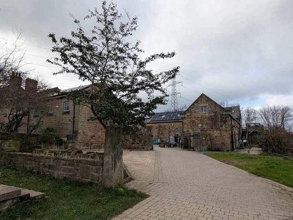 A paved courtyard with a tree in the foreground leads to rustic stone buildings with solar panels under a cloudy sky