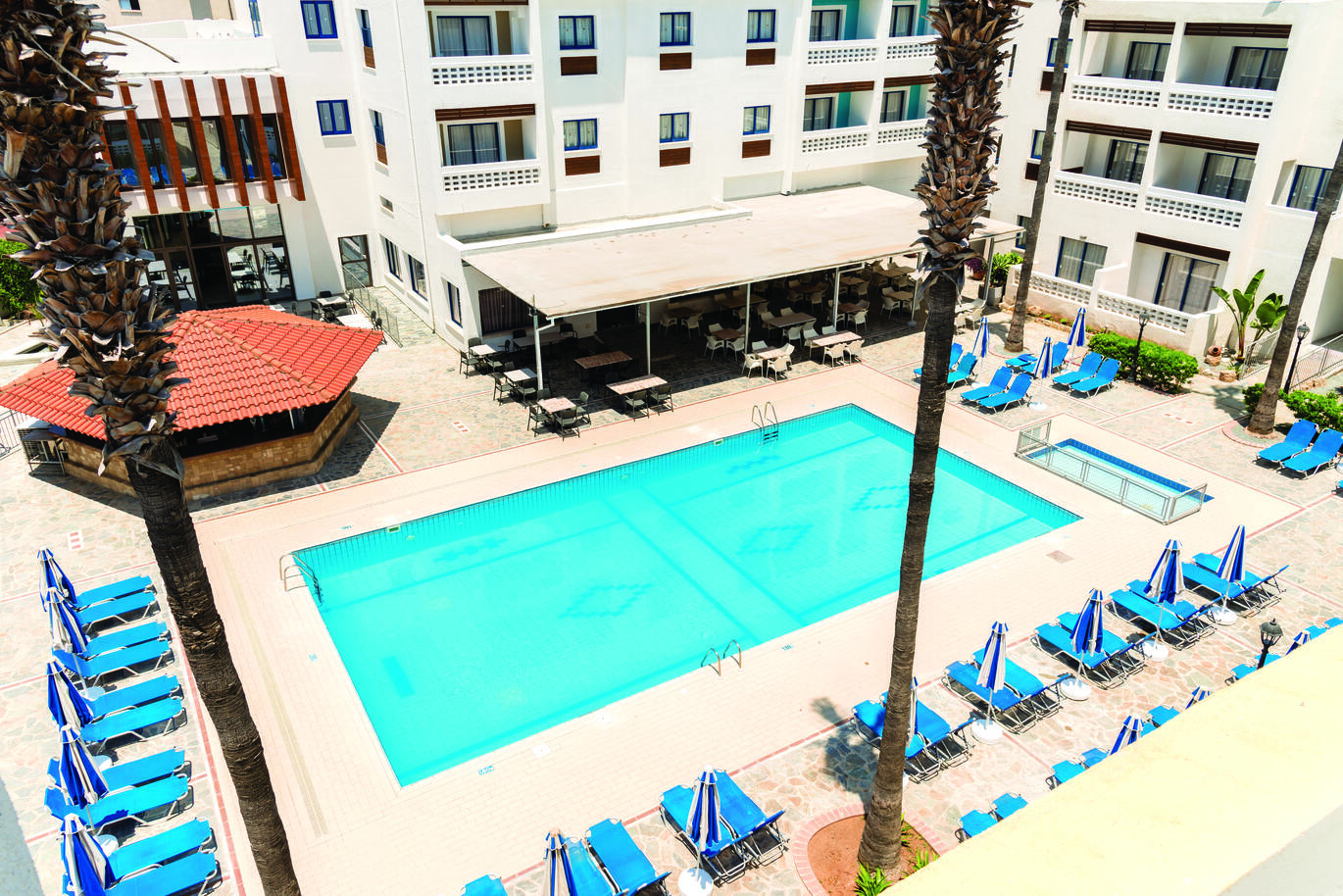 Aerial view of the pool area with sun loungers and umbrellas around. With the hotel buildings surrounding and a small undercover seating area coming off the building beside the pool.