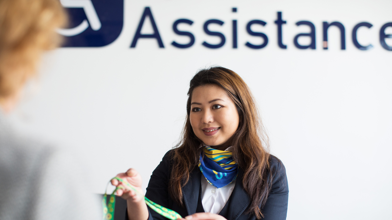 "A photo of Bristol Airport Special Assistance desk. A person in airport uniform with a green and blue scarf, blue jacket, white shirt. The word Assistance is written on the wall behind them'."