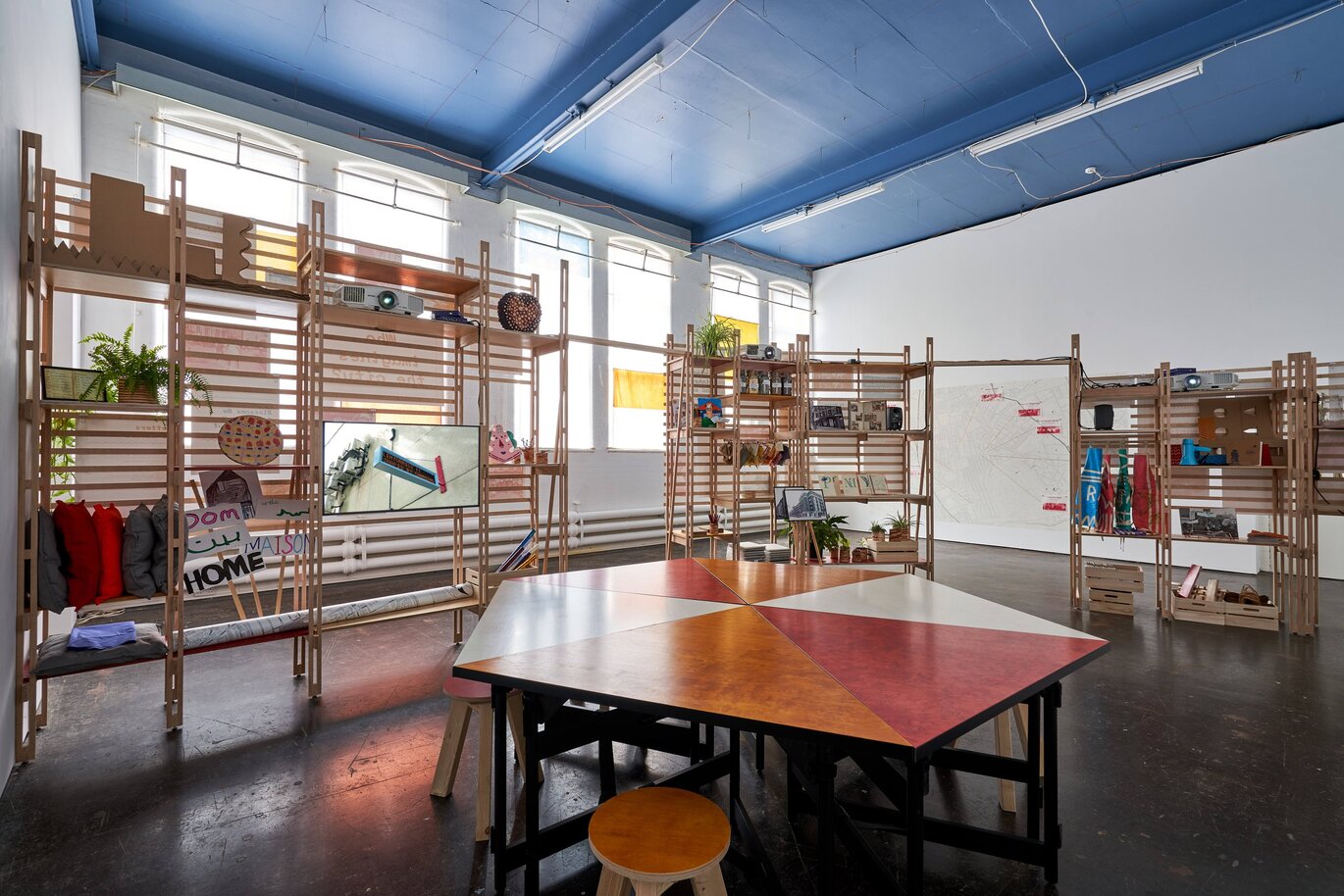View inside a room with a hexagon shaped table with round stools around it, shelves behind with various items on with windows behind and a blue ceiling above.