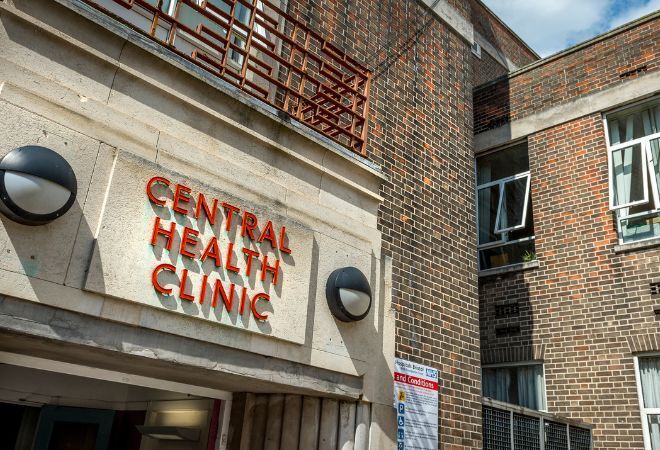 A picture of the sign above the entrance that reads 'CENTRAL HEALTH CLINIC' in red capital letters against a block cement background with a wall light either side. The building is made from brick and you can see a couple of windows in the background.