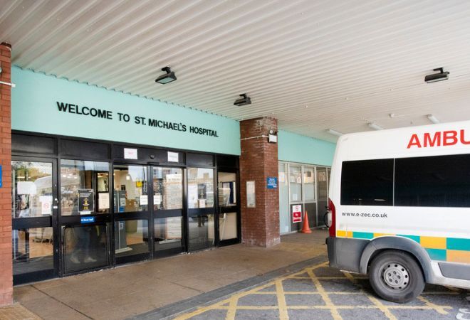 The entrance is glass doors with black borders with a light blue sign above with black letters that read 'Welcome to St. Michael's Hospital'. The ceiling is white and grooved with a few black lights. There is an ambulance parked outside of the entrance on yellow hatched markings on the floor.