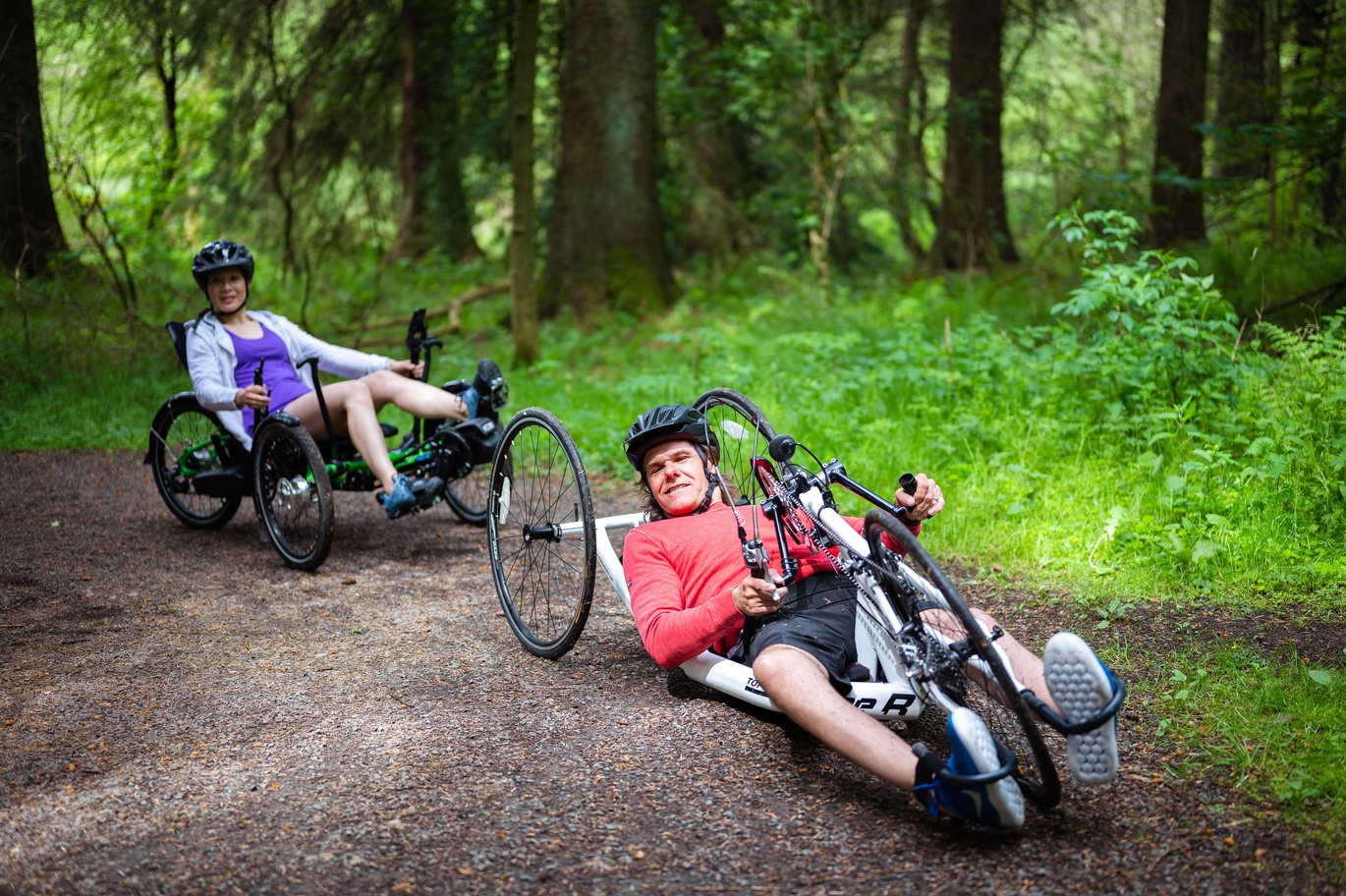 "Male and female using wheelchair cycles making way down a hill through a forest"