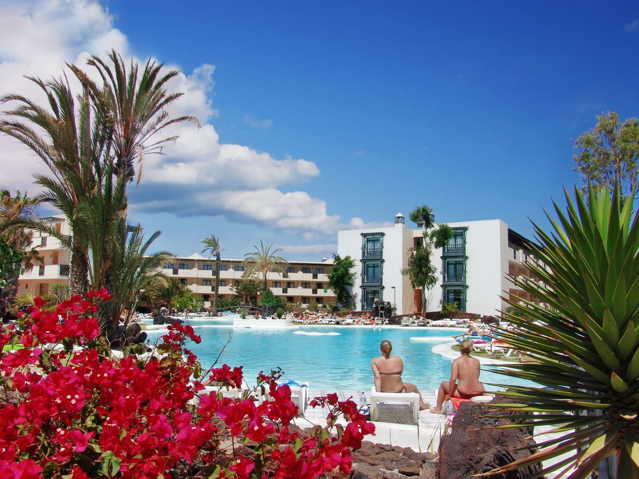 A view from behind palm trees and pink flowers of the pool and hotel buildings with people around the pool on deck chairs on a sunny, slightly cloudy day.