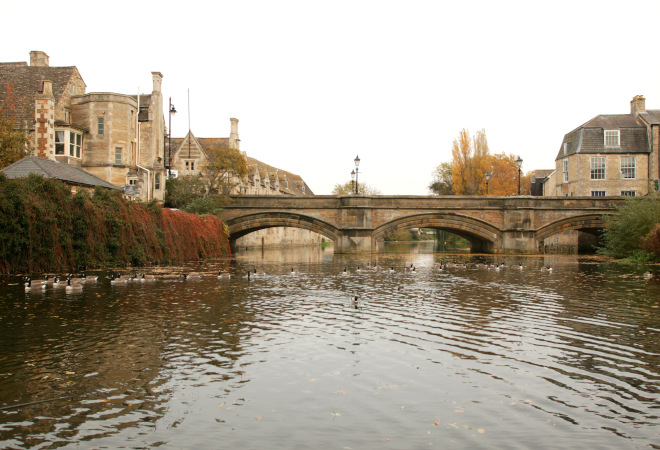 A view of a brick bridge over a river, with ducks in the water and buildings on either side