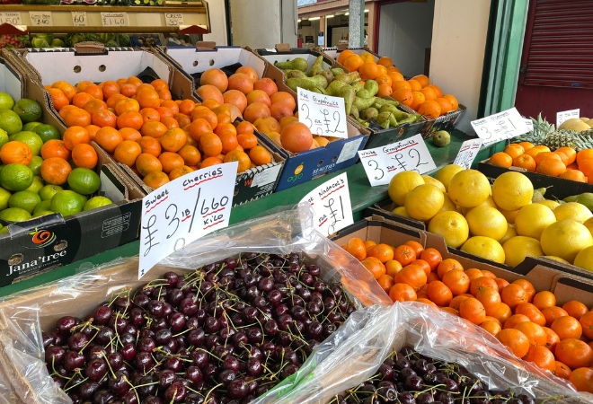 The image shows lots of different fruits in boxes - limes, oranges, grapefruit, pears, pineapple, melon and cherries with the prices above them.