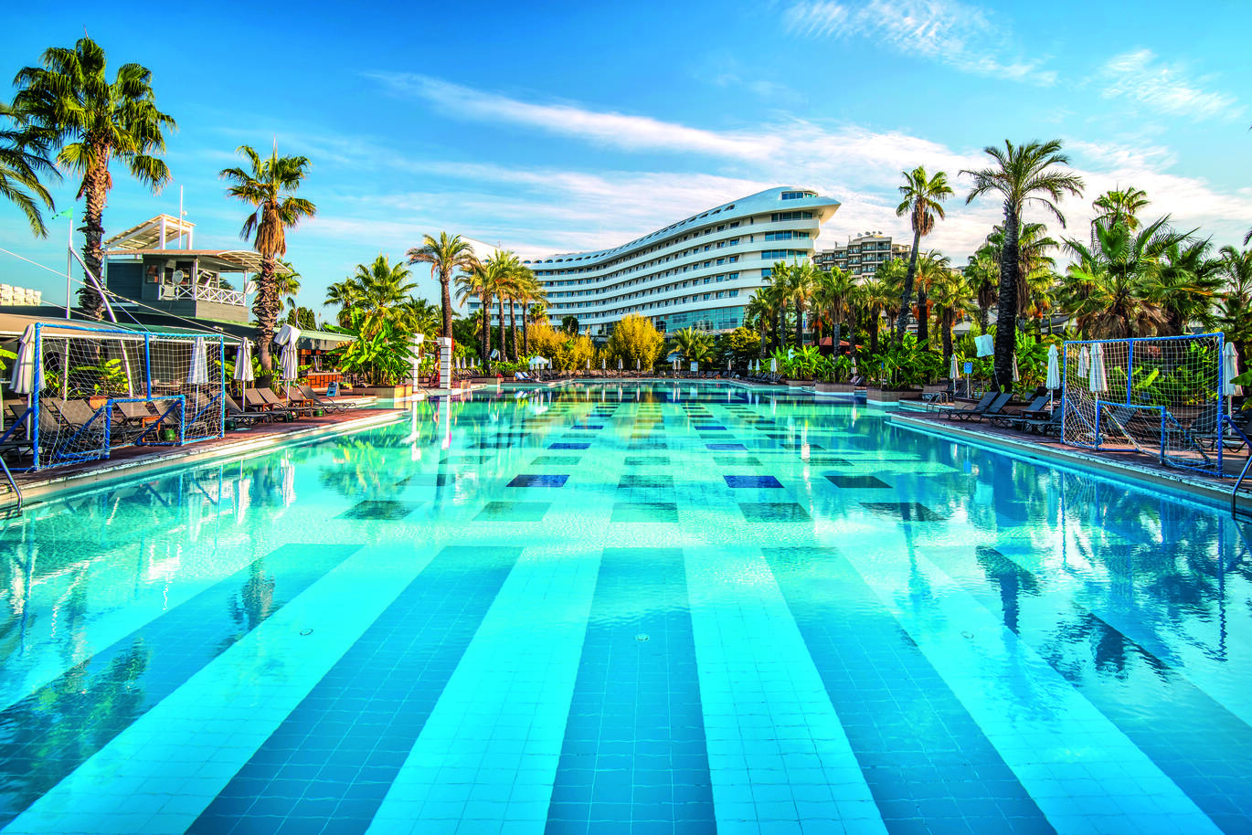 View from the large pool of the tall, boat shaped hotel building. With the pool surrounded by sun loungers, umbrellas and palm trees on a sunny day.