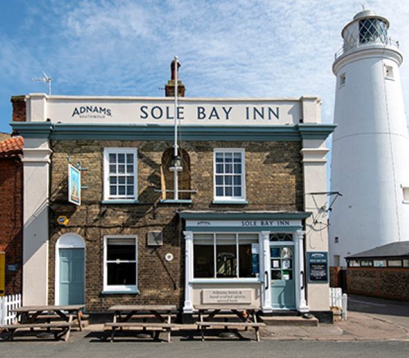 "External of The Sole Bay Inn - a brick building with blue window frames and a white lighthouse can be seen in the distance"