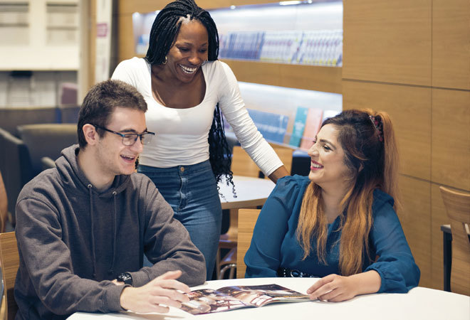 photo of two students sat at a table laughing at a magazine, with a third student stood between them also laughing.