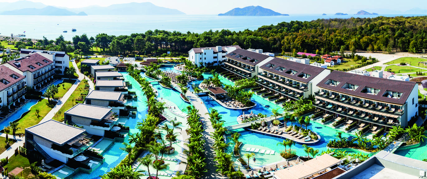 Aerial view of the clusters of hotel rooms with the swimming pool in the  middle with small islands in the middle with sun loungers on and a few trees on each. There's also a swim up cluster of rooms to the left. Behind the hotel you can see many trees with the sea and mountains in the background.
