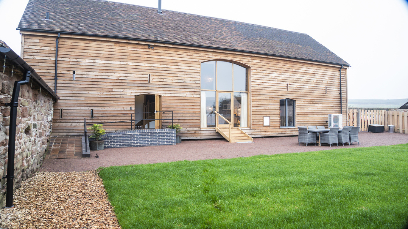 The exterior of the wooden built Sandybury Barn with a table and chairs on the patio and the lawn in front.