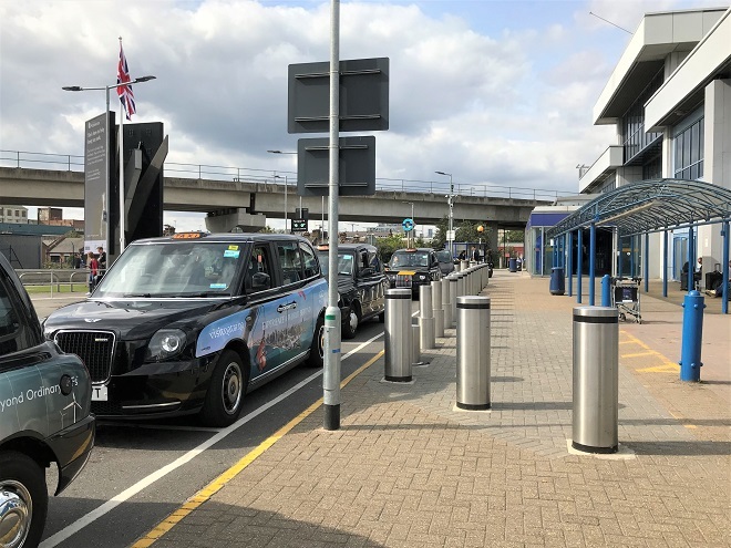 'Photo of taxi rank outside London City Airport. Grey bollards between taxis and shelter. Seating available near shelter.'