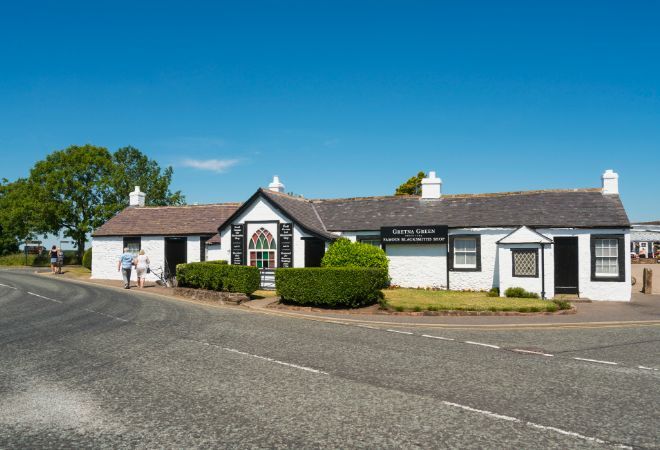 "view from across the road of the Gretna Green Famous Blacksmiths. A white cottage with a black roof, bushes in front. A two sets of couples are walking past the building"