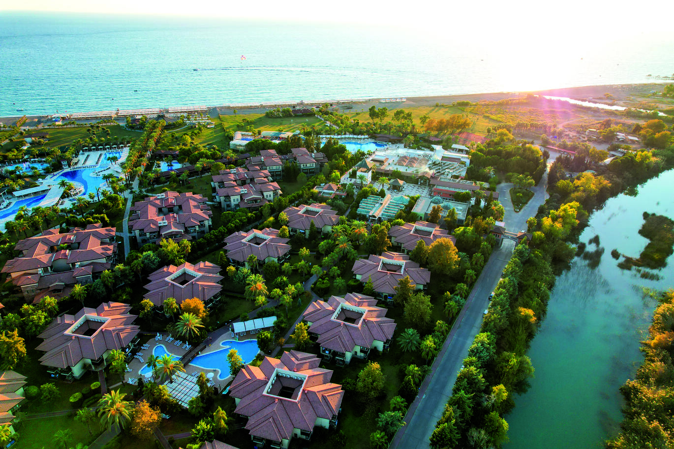 Aerial view of the hotel complex with clusters of villas and pools dotted around with many trees surrounding. There is a river right behind and the beach and sea in front.