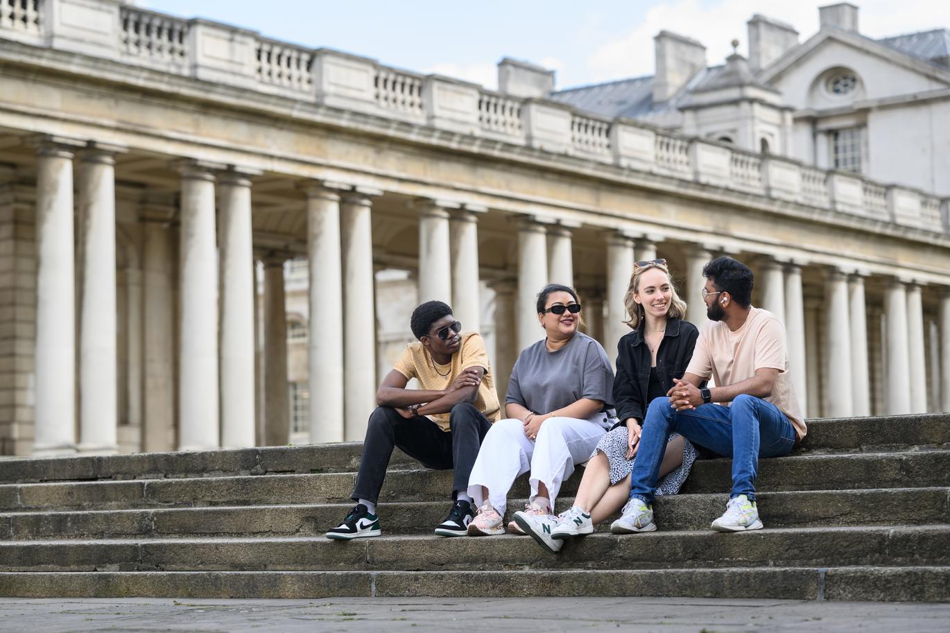 "Five students sit together on stone steps in front of a historic building with tall columns, talking as a group. The scene is outdoors on a bright day, with classical architecture filling the background"