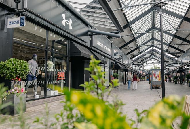 "Covered outdoor shopping walkway with Under Armour store and sale sign, lined with plants and benches"