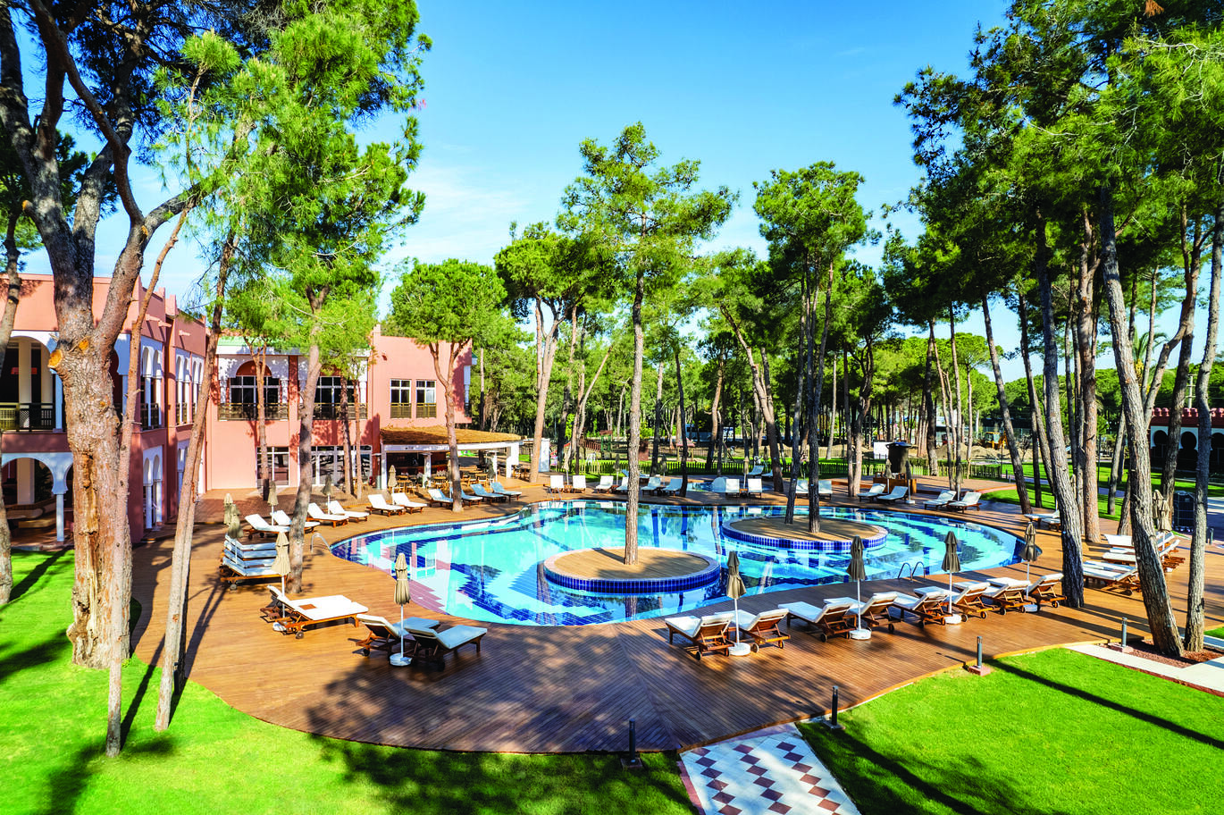 View of the pool area with sun loungers and umbrellas around the wooden decking of the pool and lawns behind, with two small islands in the pool with tall trees on them and many trees around the area. With a small part of the hotel building visible to the left and the blue sky above visible through the trees.