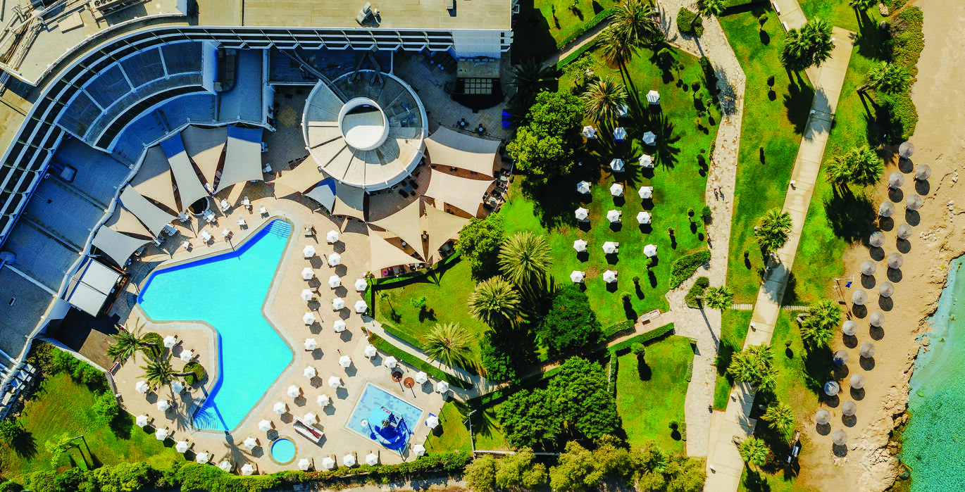 Birdseye view of the pool area with lots of greenery then leading to the beach and sea behind, with the hotel building surrounding the pool and surrounded by deck chairs and umbrellas.