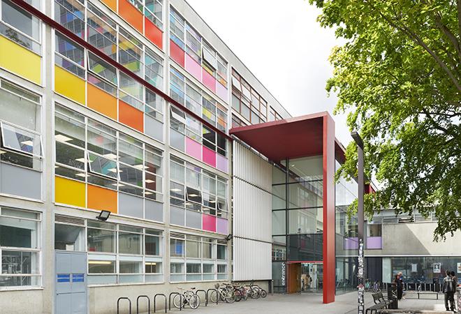 “Modern building with large glass entrance framed by a red structure, and exterior walls featuring rows of colorful panels in orange, yellow, pink, and gray. Several bicycles are parked near the entrance, and a leafy tree partially shades the foreground.”