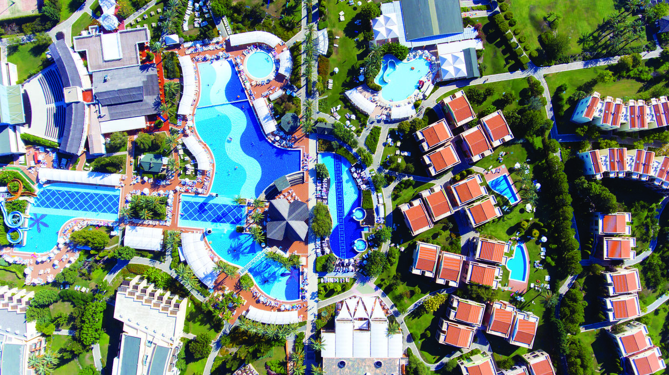 Birdseye view of the hotel complex with lots of buildings and 4 main pools all within lots of green grass and trees. 
