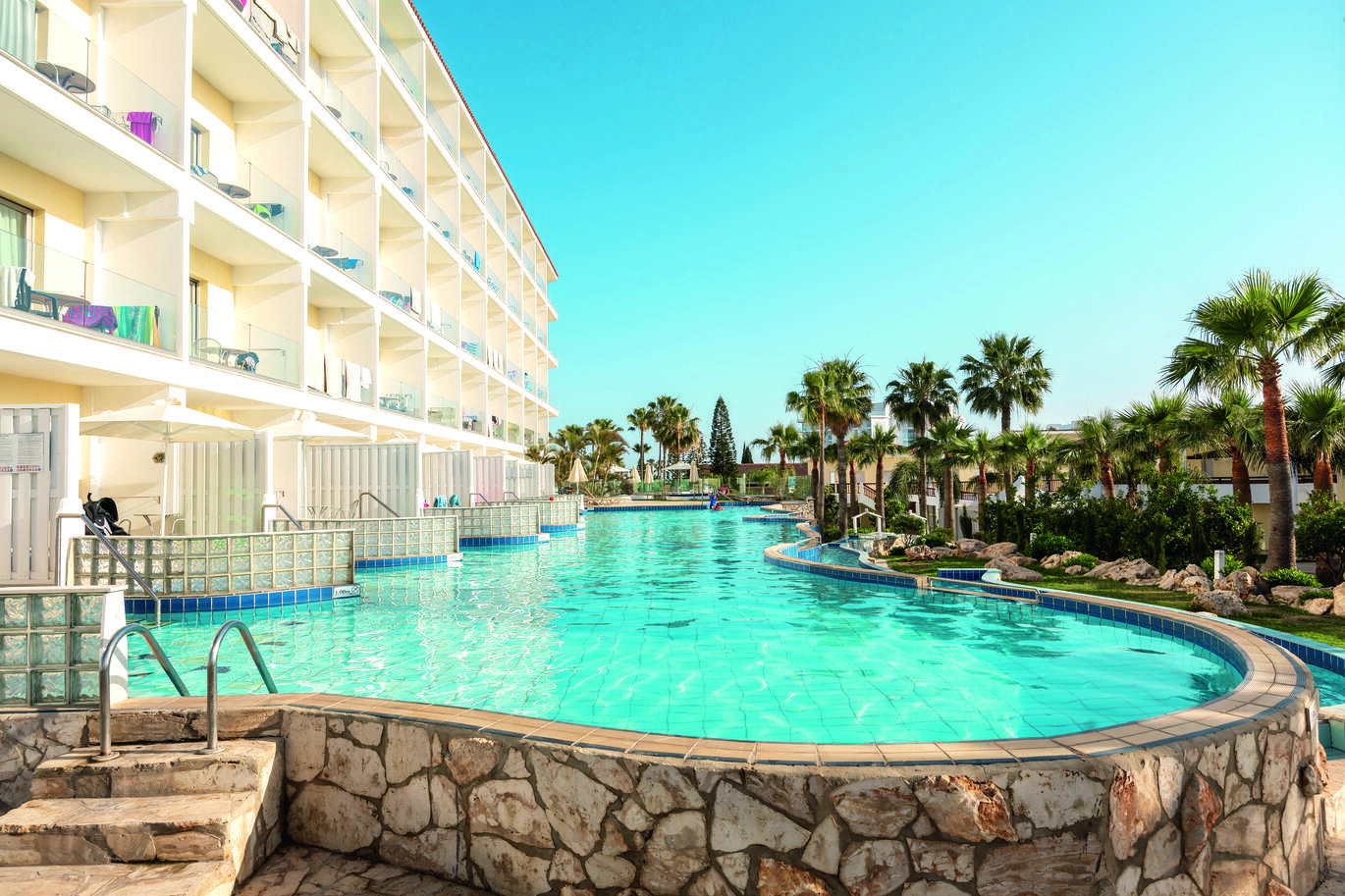 A long, clear blue pool surrounded by a stone wall with steps into it in front of a cluster of hotel rooms. The pool is surrounded by palm trees and the sky above is clear and blue.