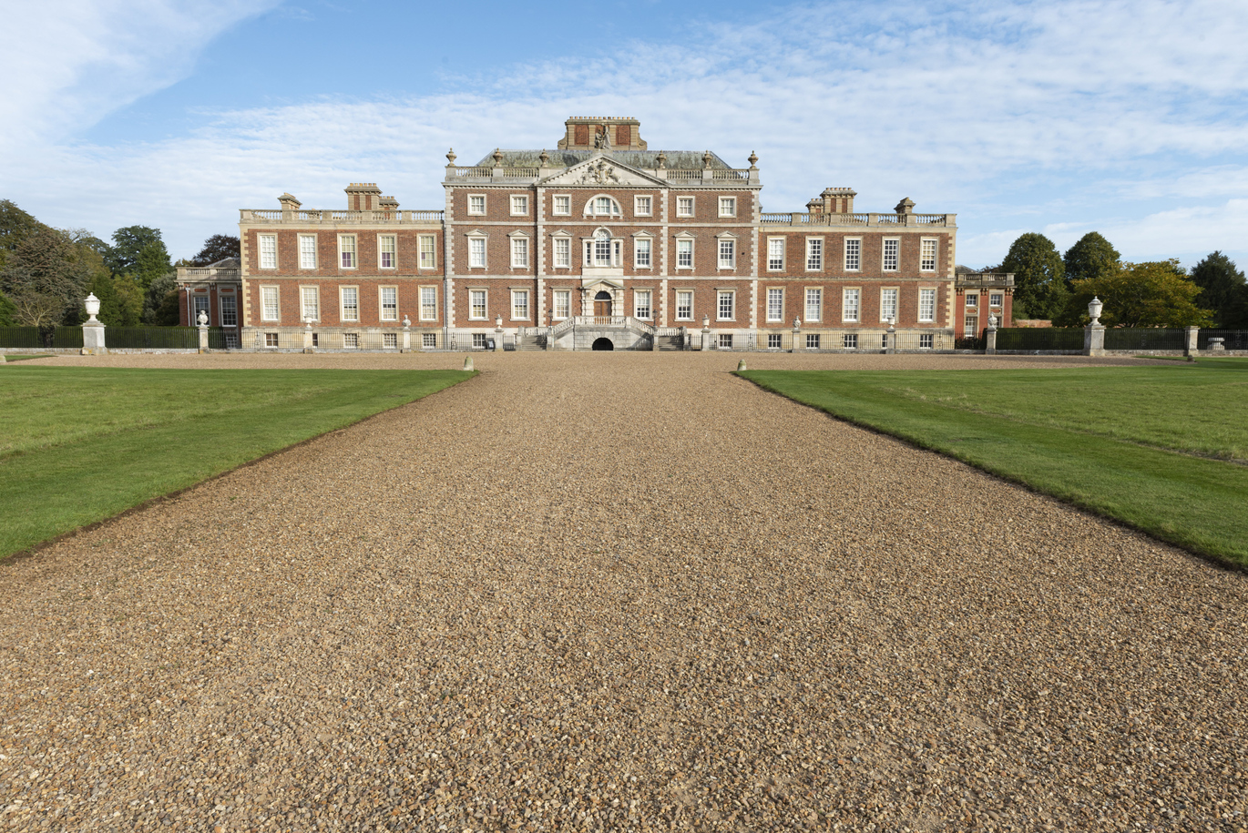 "The south front of Wimpole Hall in Cambridgeshire. It is a large, grand brick-built 17th century mansion with a large gravel driveway and well-maintained lawn in the foreground".