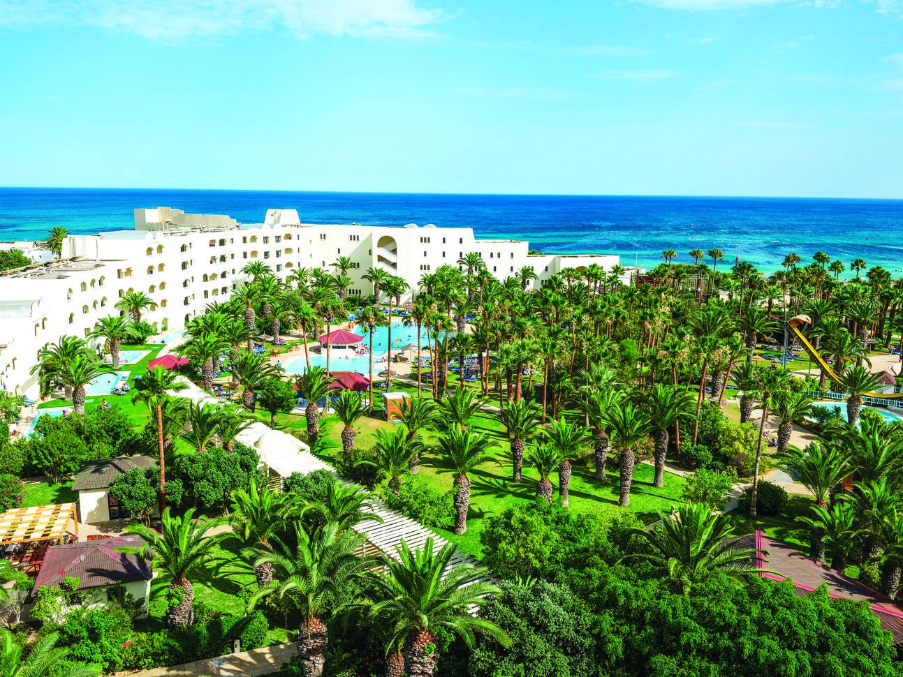 Aerial view of the hotel complex with pools dotted around surrounded by green lawns and many palm trees with the deep blue sea visible in the background and blue sky above.