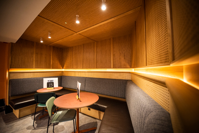 : Interior of a modern cafe with warm ambient lighting. The space features a cozy corner booth with dark upholstered seating and a round wooden table. The walls are lined with geometrically patterned wood panels, and the ceiling has recessed lighting. 