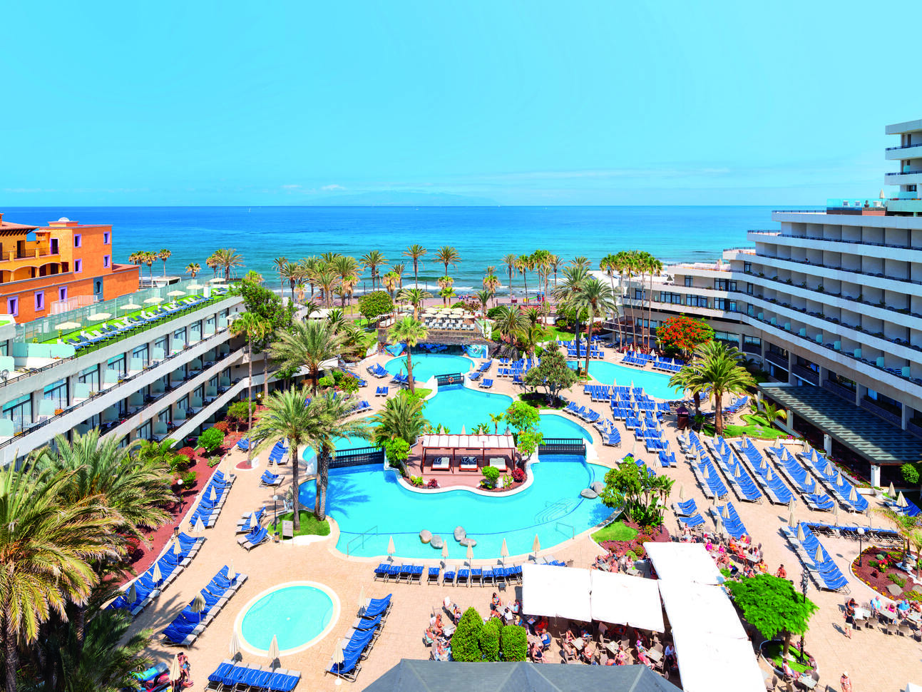 Aerial view of the large pool area surrounded by sun loungers, a small island in the middle of the pool with sheltered sunbeds with bridges either side and a small bridge over the pool further up. The pool area is surrounded by the hotel buildings with palm trees dotted around and the beach an sea visible behind with the blue sky above.