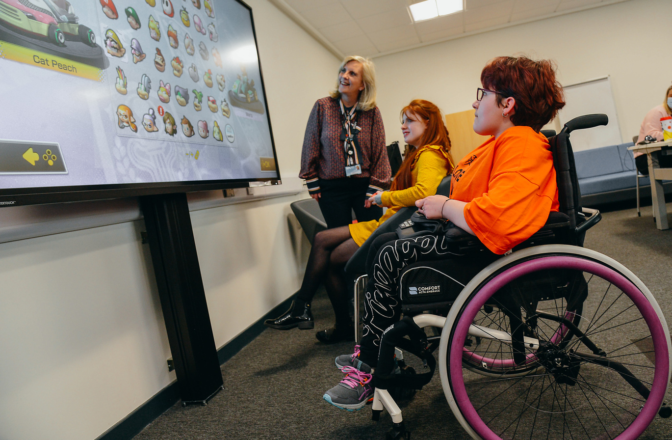 "Group of students and a teacher, looking at a large digital screen. One is in a wheelchair, one sitting on a seat and one standing"