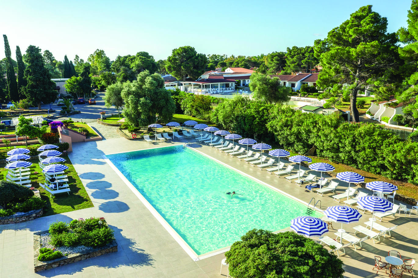 View of the pool area with a single person swimming surrounded by sun beds with blue and white striped umbrellas. There are many trees and greenery around with more of the hotel buildings visible in the background.