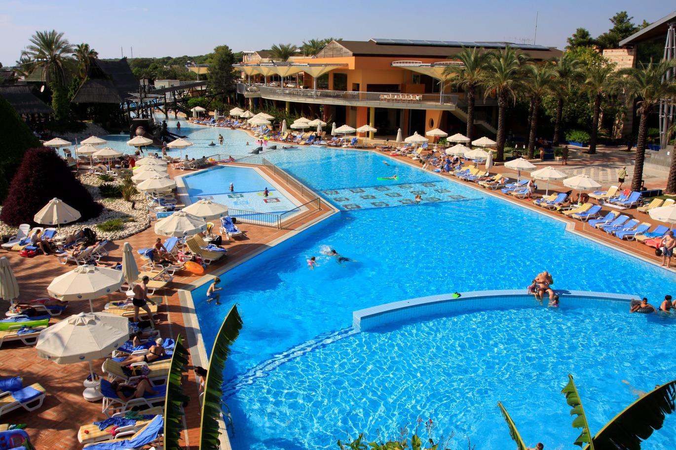 A large swimming pool surrounded by blue sun loungers and white umbrellas with a number of people in the pool and around the pool. There are also a number of trees surrounding the pool and hotel building which is further back in the image, next to the pool. It is a lovely clear day with a blue sky.