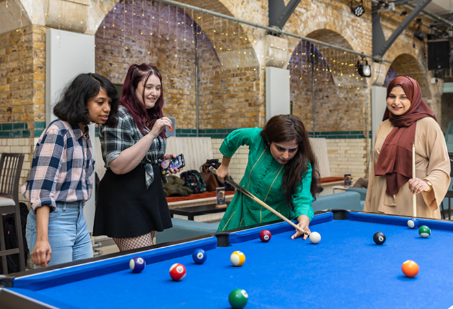 "Image of four female presenting students playing pool inside of a CCCU building"