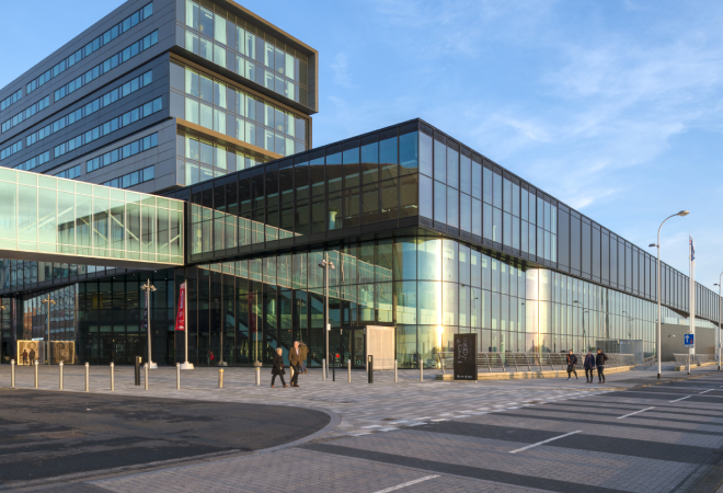 "Corner of a building with multiple glass windows, with a glass bridge walkway on the left directing towards another building out of view"