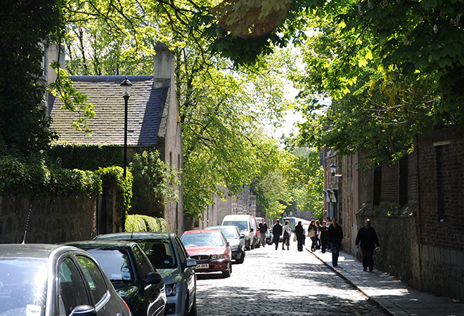 "Street view of parked cars and pedestrians walking on pavement and road lined with trees"