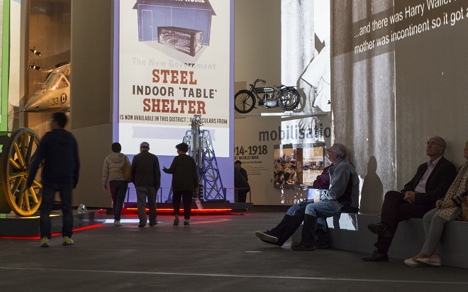 Inside an exhibit at IWM North. People are browsing the exhibit. There is an image projected of a Steel Indoor Table Shelter with this wording underneath.