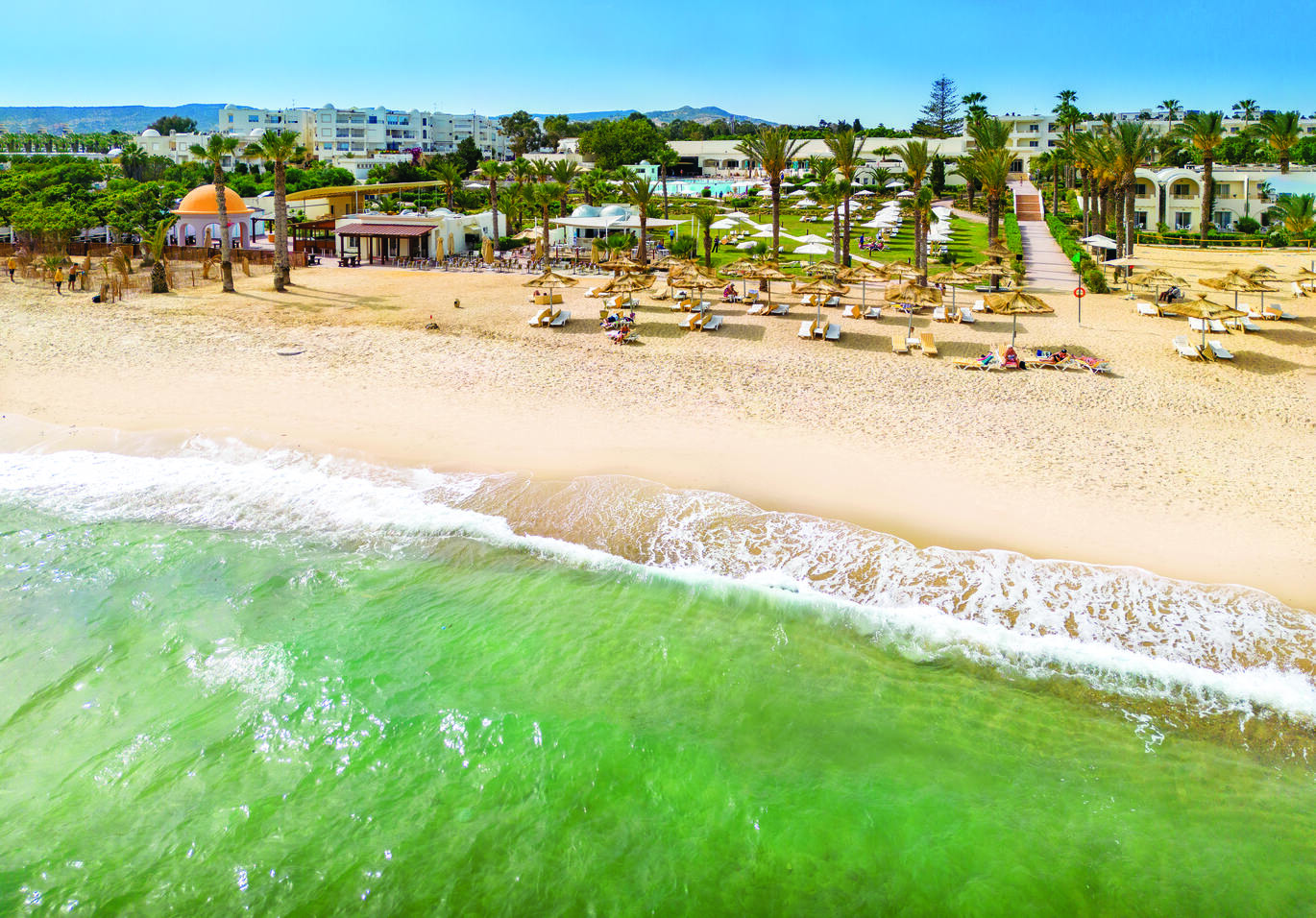 Aerial view of the beach and sea with sun loungers on the beach and the hotel complex behind.