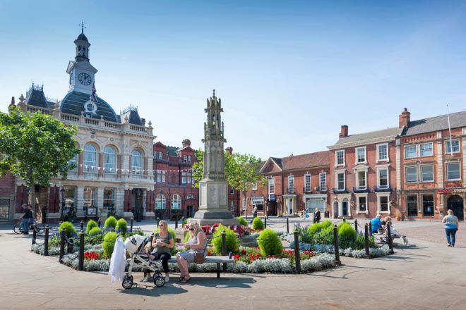 "photo of Retford Market Square. the town hall is in the background, with a clock on the roof. a cenotaph is centre, with flowers growing around it. two people sit on a bench in the foreground, with a baby."